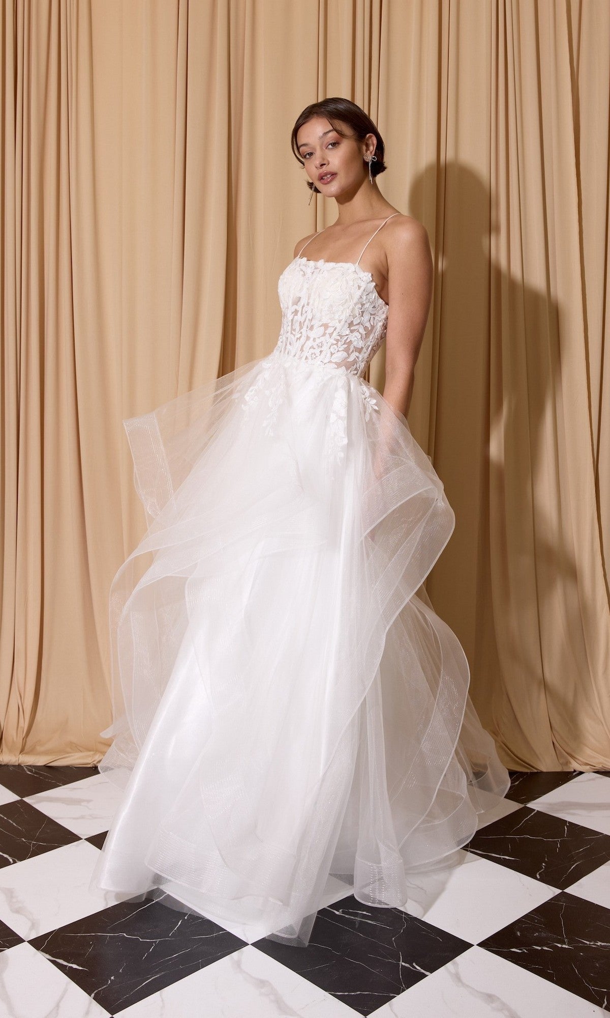 A woman stands on a black-and-white checkered floor in front of beige curtains, wearing the Eureka EK167 Long Prom Dress—a white strapless gown with a floral lace embroidered corset and layered tulle A-line skirt.